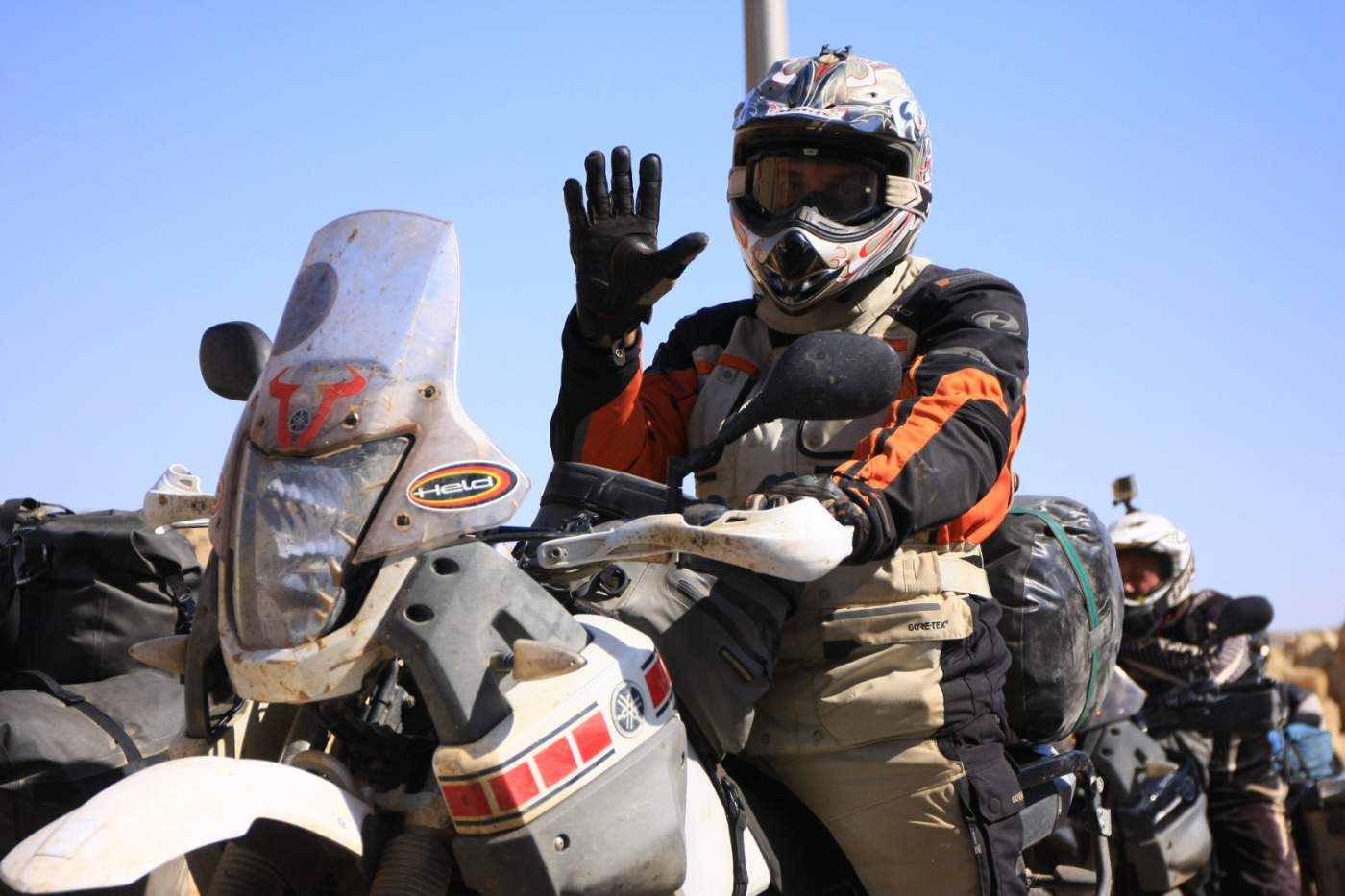 Motorradfahrer in Reisekleidung auf beladener Reiseenduro in Afrika, Hand zum Gruß erhoben