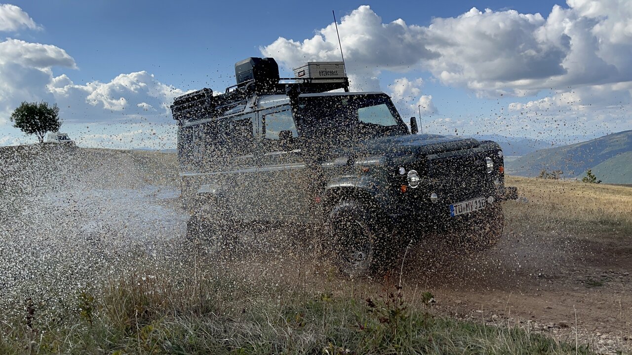 Ein schwarzer Land Rover Defender durchquert platschend Schlamm und Wasser auf einem Hangweg