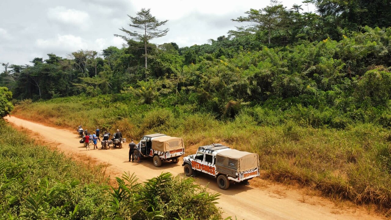 Two Overcross zebra-patterned trucks and a group with motorcycles travel a red dirt track through dense jungle