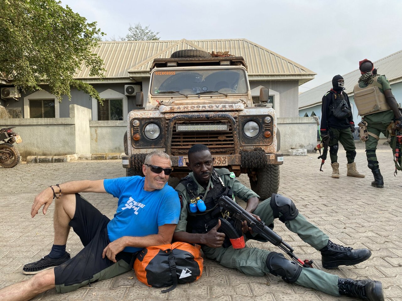 Um viajante senta-se ao lado de uma escolta armada em frente ao Land Rover de expedição, com soldados por perto