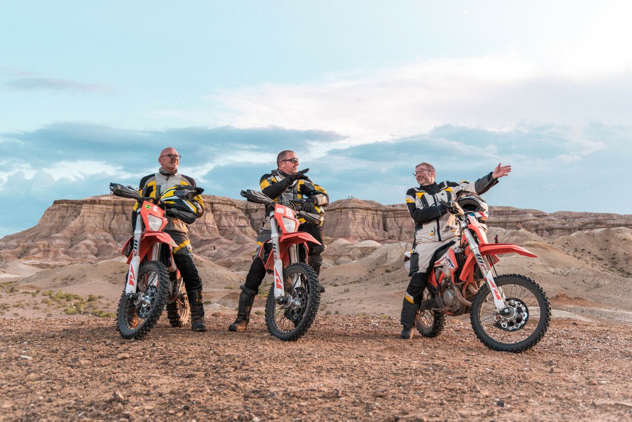 Drei Fahrer mit Enduro-Motorrädern stehen in der Abenddämmerung vor gestreiften Badlands-Klippen