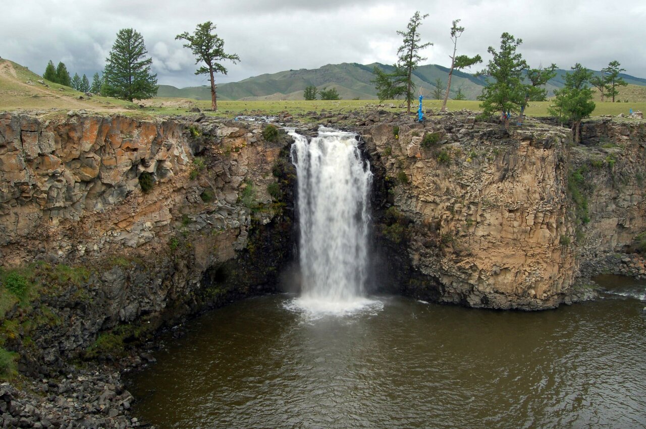 Ein Wasserfall stürzt über Basaltfelsen in einen Fluss, an nahen Bäumen hängen Gebetsbänder