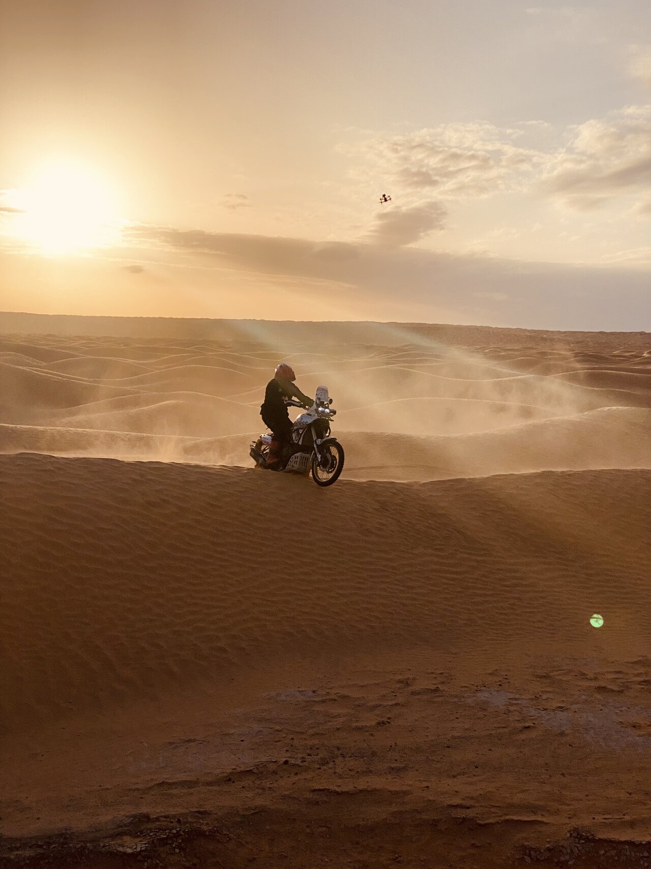 Um motociclista percorre a crista de uma duna ao pôr do sol enquanto um drone filma do alto