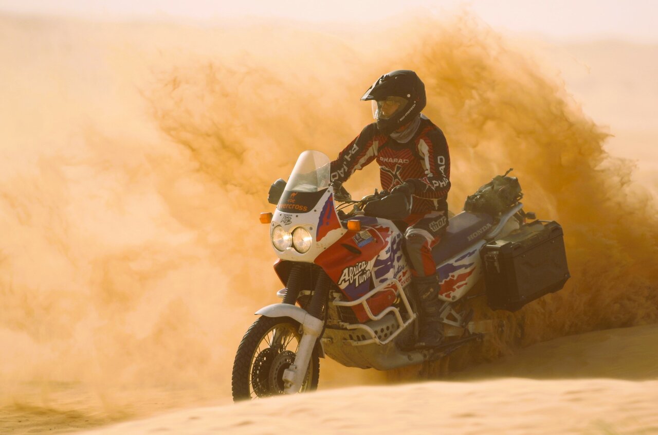 A rider on a Honda Africa Twin motorcycle sprays orange sand while cresting a Sahara dune