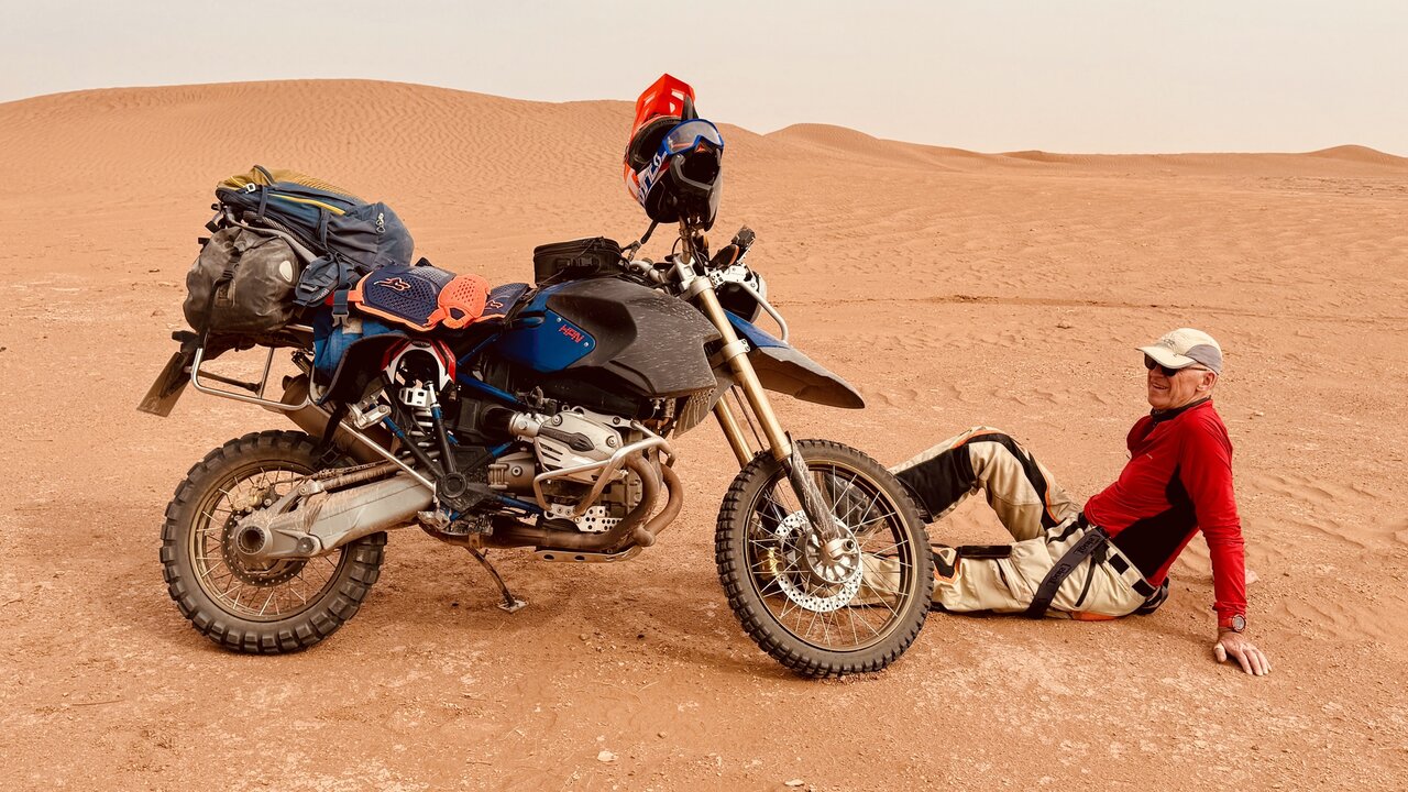 Ein schwer beladenes BMW-Enduro-Motorrad, daneben ruht ein Fahrer in den Dünen