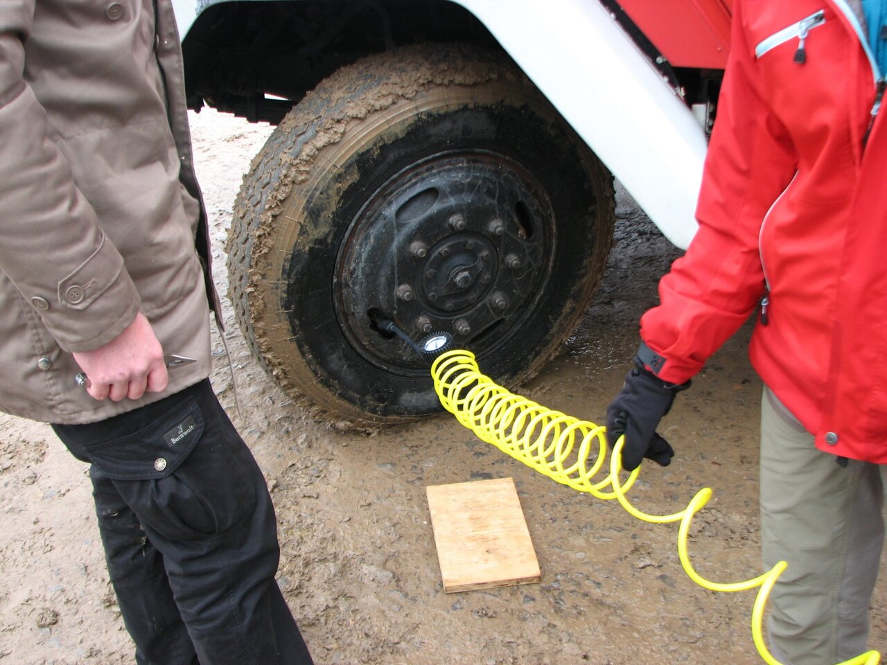 Two people connecting a yellow coiled air hose to a truck wheel on muddy ground