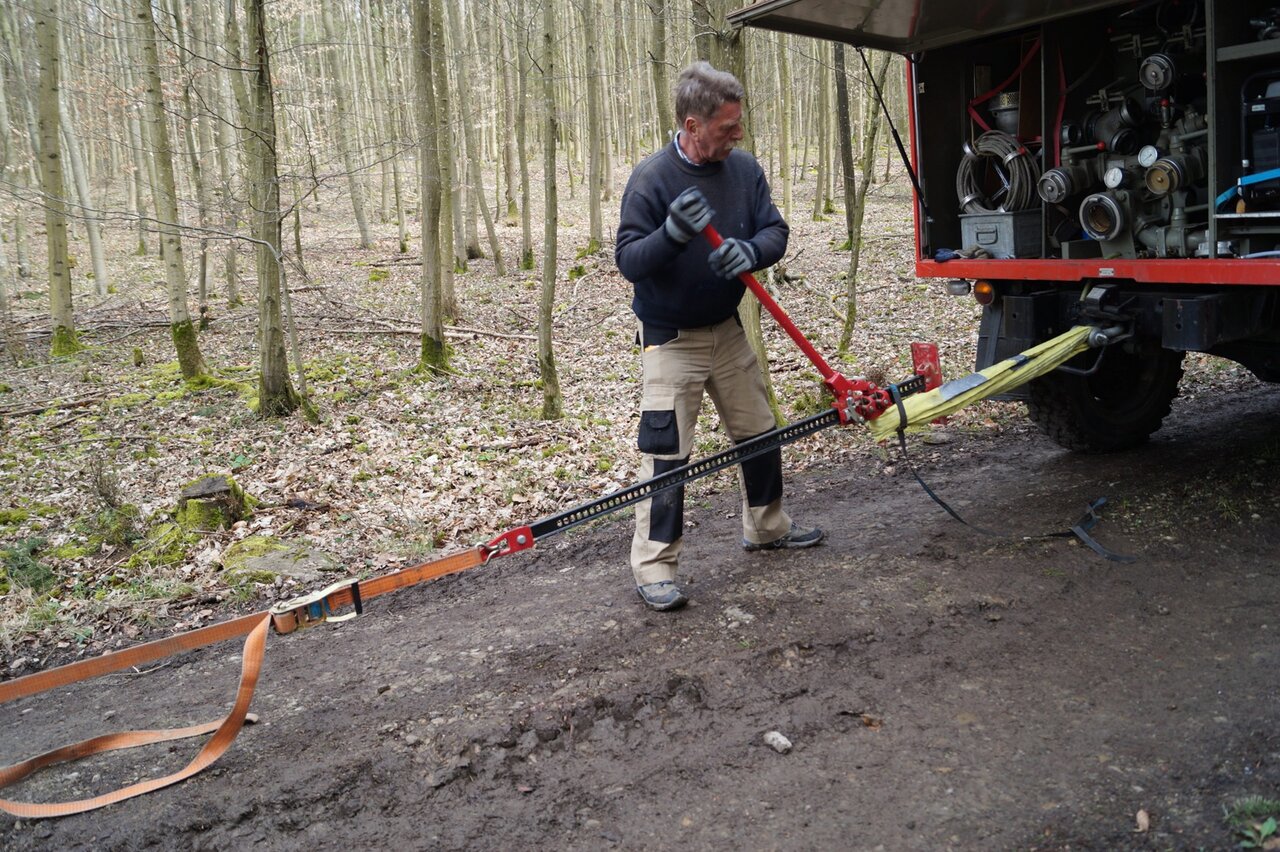 A man operating a ratchet strap tensioner behind a red service truck in a forest