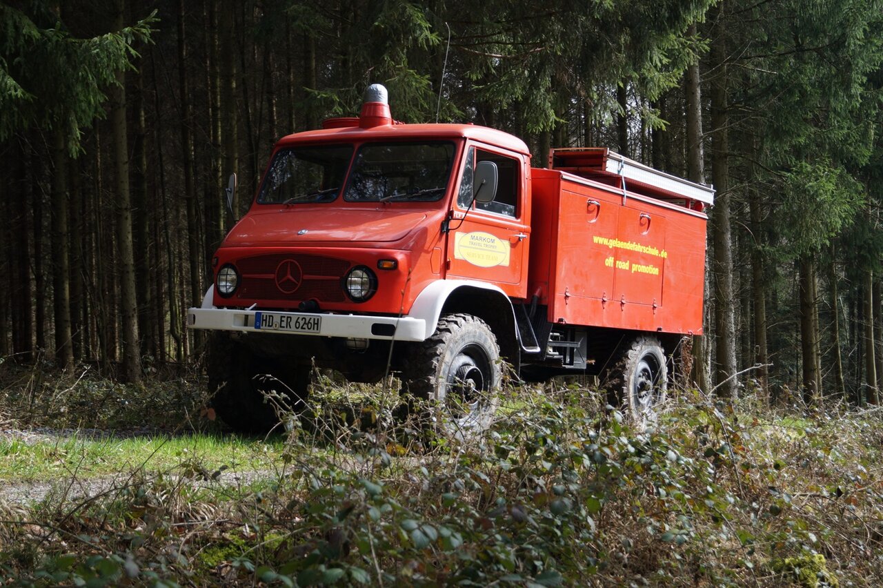 A red Mercedes Unimog service truck marked Markom Travel Trophy, parked in a forest