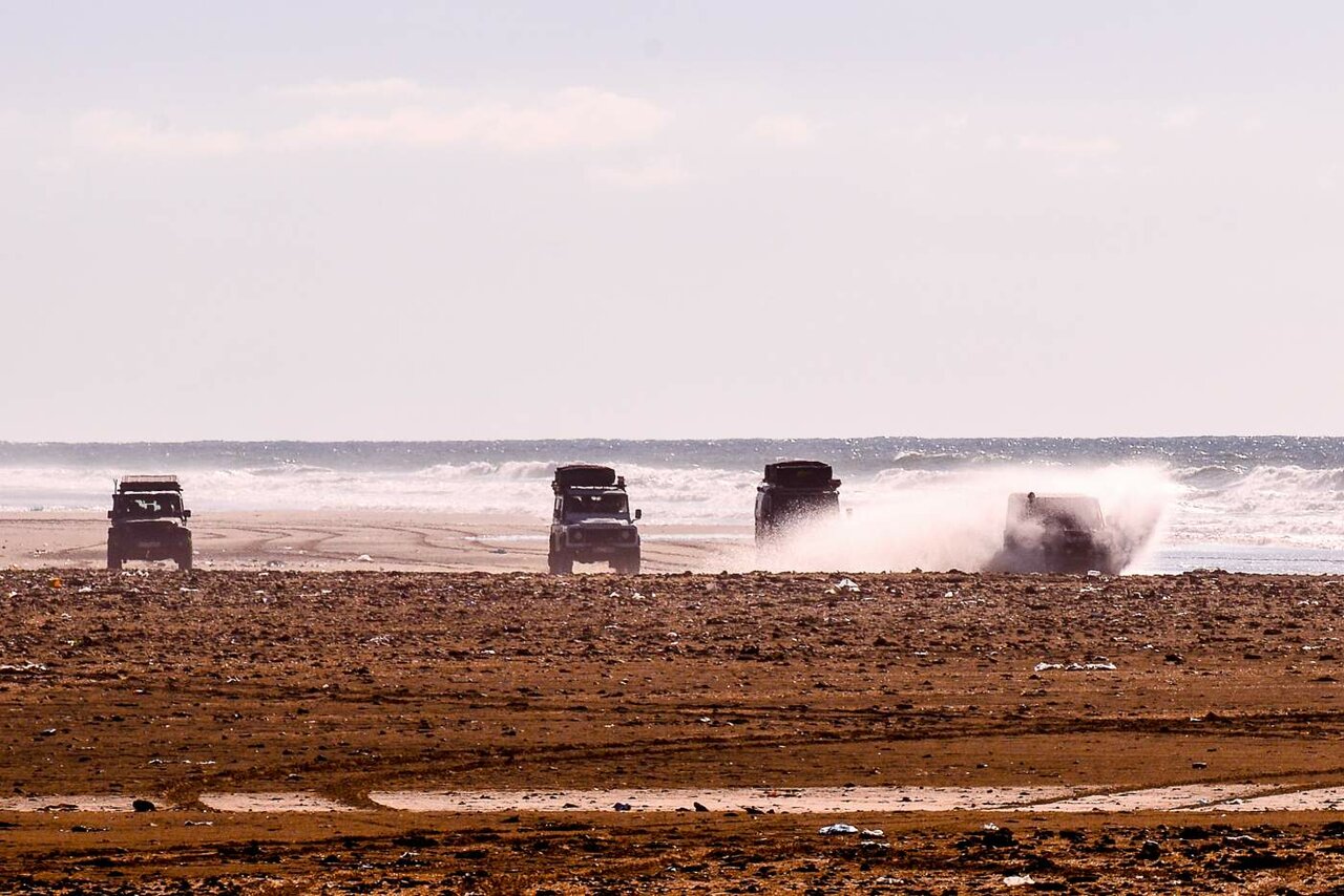 Vier Land Rover Defender fahren am Strand entlang, einer durchquert spritzend die Brandung