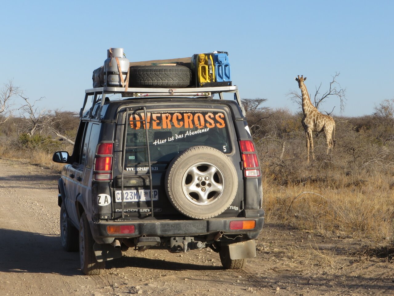 Ein Land Rover parkt auf einer Schotterpiste, in der Nähe grast eine Giraffe
