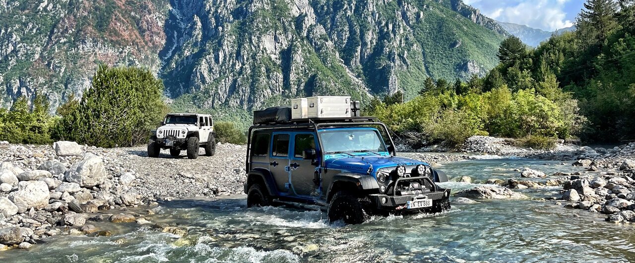 Dois Jeep Wranglers atravessam um rio pouco profundo num vale rochoso íngreme