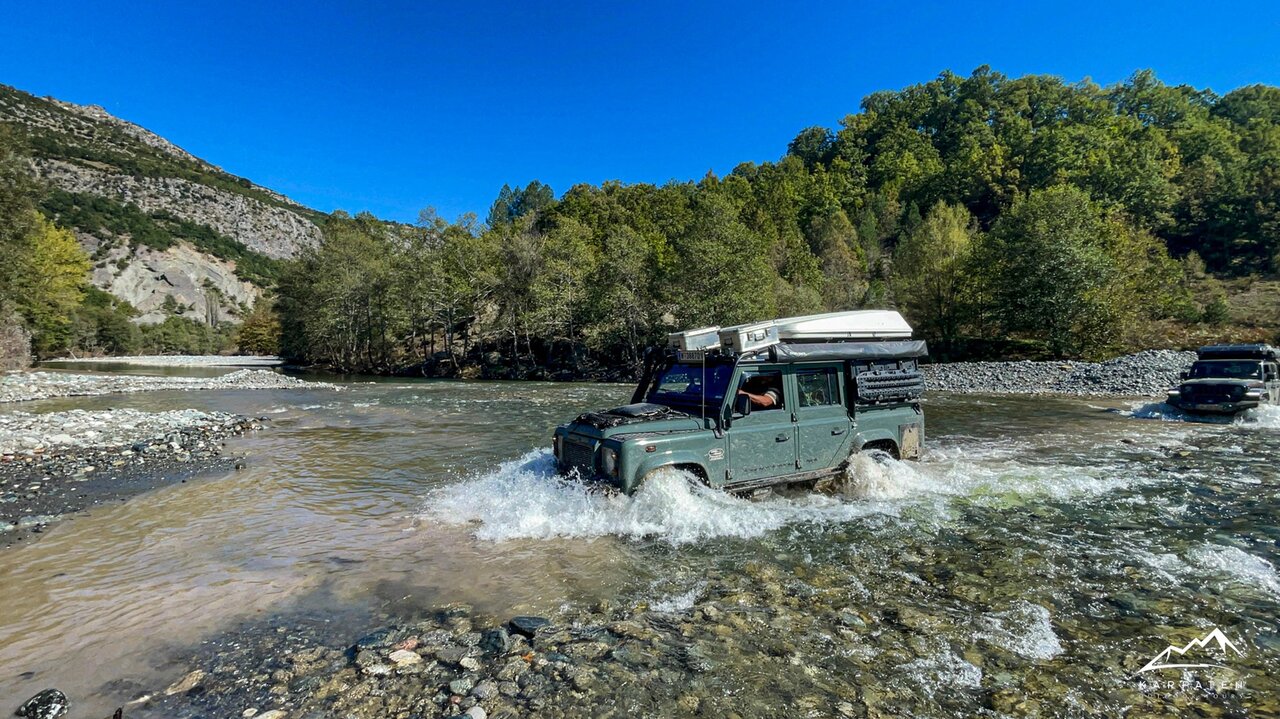 Um 4x4 com tenda no tejadilho atravessa um rio raso perto de colinas verdes