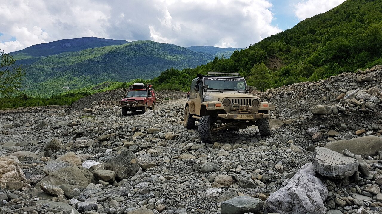 Ein beiger Jeep und ein roter SUV durchqueren ein trockenes, steiniges Flussbett unter grünen bewaldeten Bergen