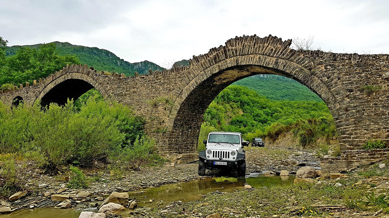 Ein weißer Jeep und weitere Geländewagen durchqueren einen Fluss unter einer alten steinernen Bogenbrücke