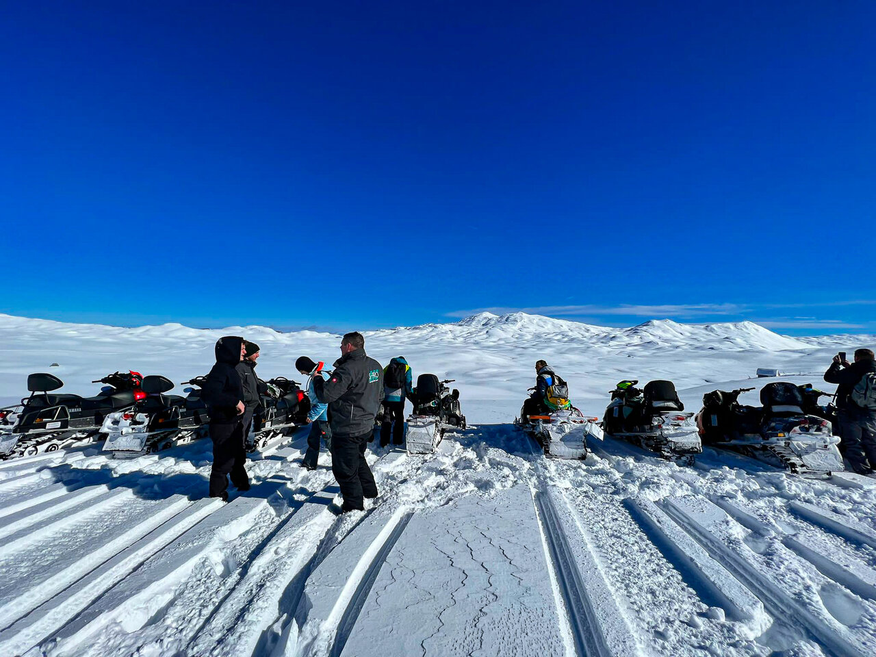 A group of riders and snowmobiles gathered on a sunny snowy plateau