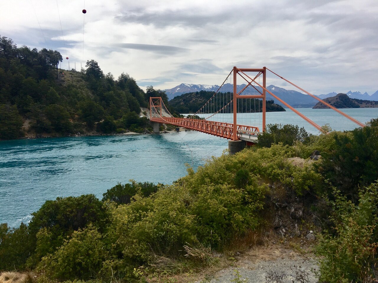 An orange suspension bridge crossing a turquoise river with mountains behind
