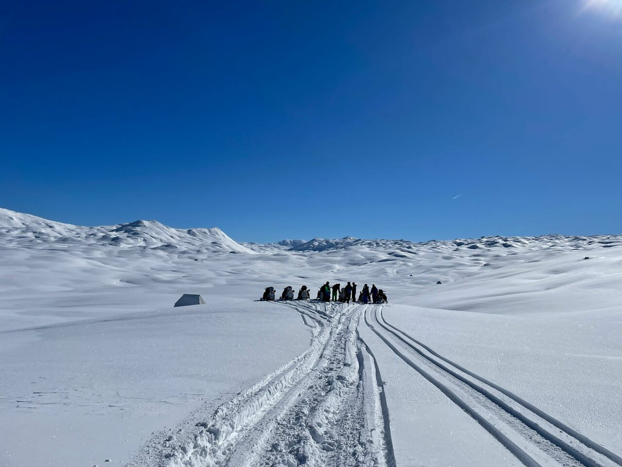 Snowmobiles and riders in the distance beside a tent on a snowy plateau