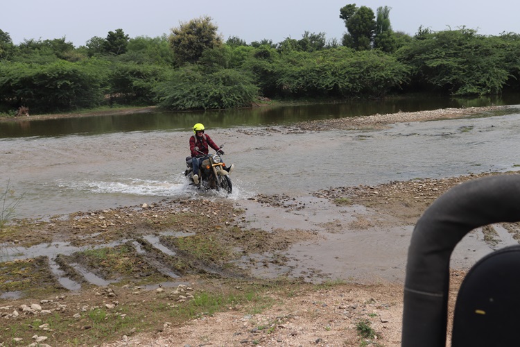 Un motorista salpica al cruzar un río poco profundo sobre barro y piedras