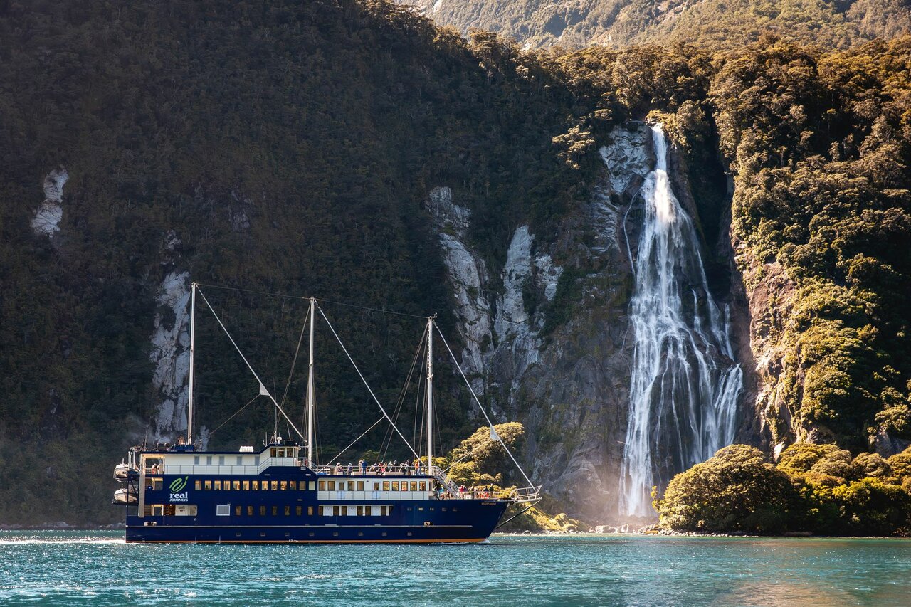 Ein Kreuzfahrtschiff nahe einem hohen Wasserfall an bewaldeten Klippen in einem Fjord