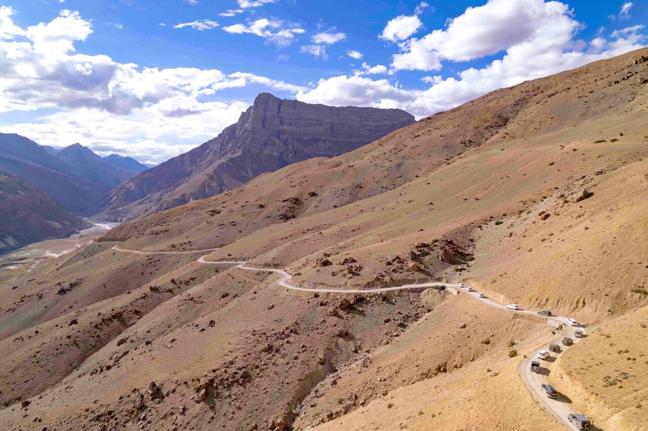 Eine kurvenreiche Schotterstraße mit einer Autoschlange, die ein trockenes Himalaya-Tal hinabfährt