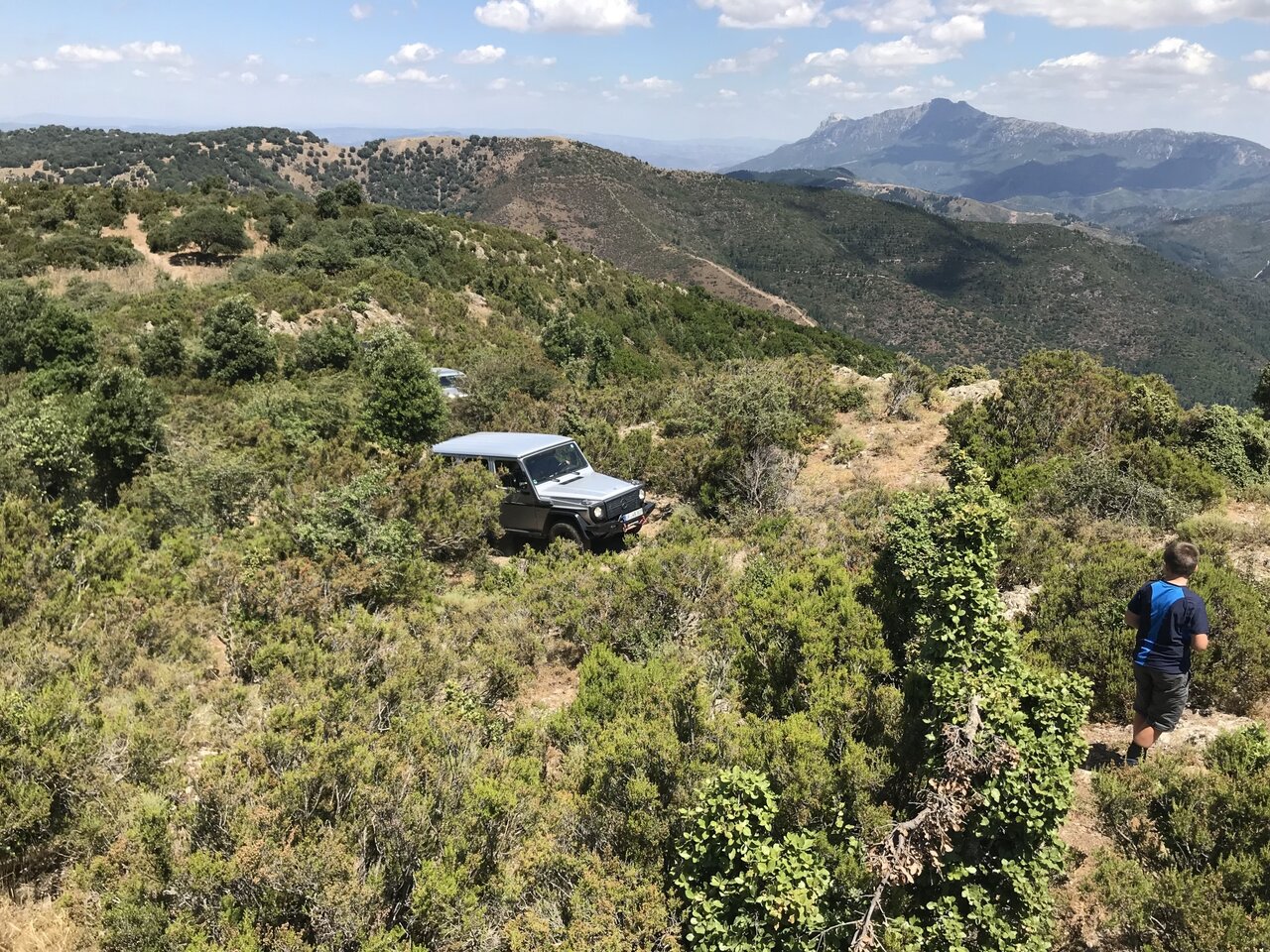 Silver Mercedes G-Class off-road vehicle on a scrub covered ridge with a hiker looking over the valley