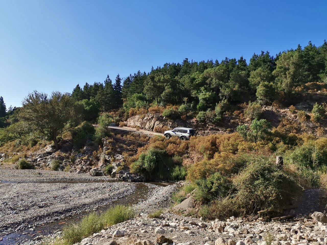 White off-road vehicle descending a gravel forest road toward a dry riverbed, seen from above