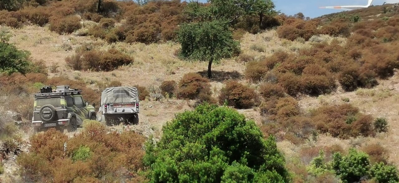 Two off-road vehicles among dry scrub on a hillside, a wing tip visible in the corner