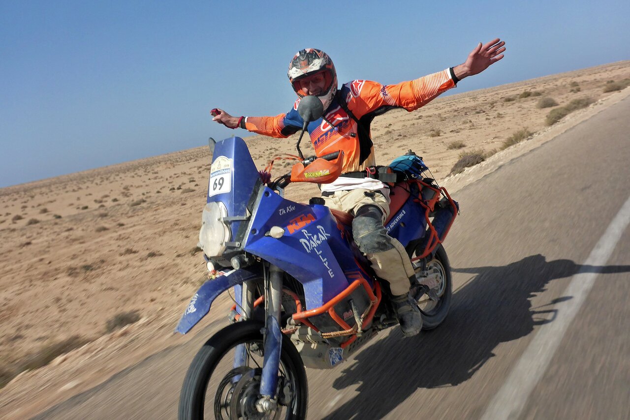 Ein Motorradfahrer streckt beide Arme hoch, während er ein Dakar-lackiertes Motorrad über eine Wüstenstraße fährt