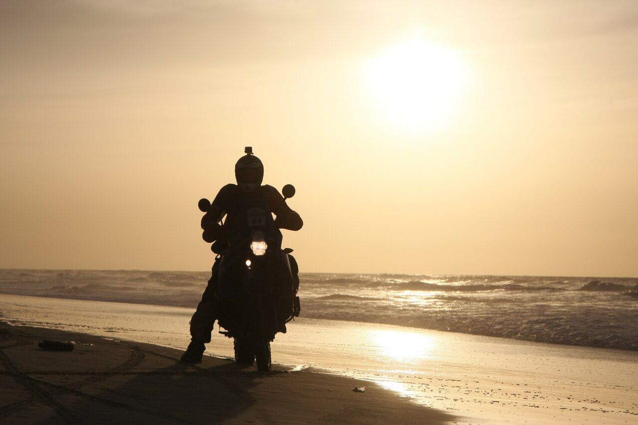 Ein Motorradfahrer ist bei Sonnenuntergang als Silhouette an einem Strand zu sehen, Meereswellen dahinter