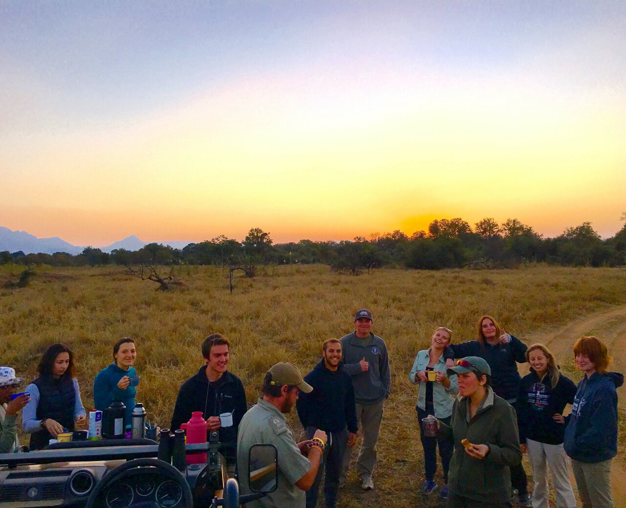 Eine Gruppe teilt sich Kaffeetassen bei Sonnenuntergang neben einem Safarifahrzeug