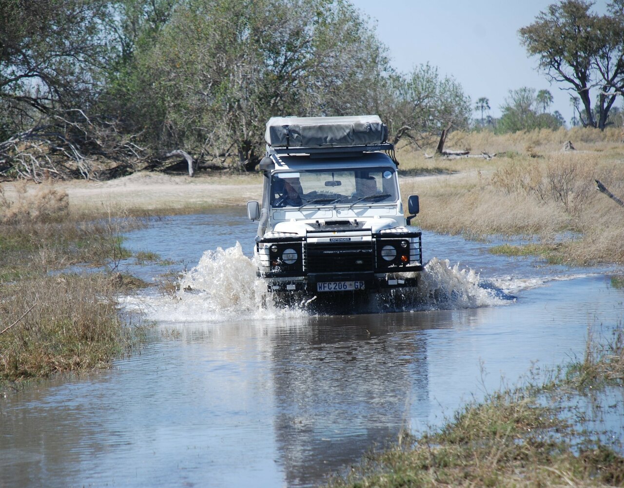 Un Land Rover Defender salpica al cruzar una llanura de hierba inundada bordeada de árboles