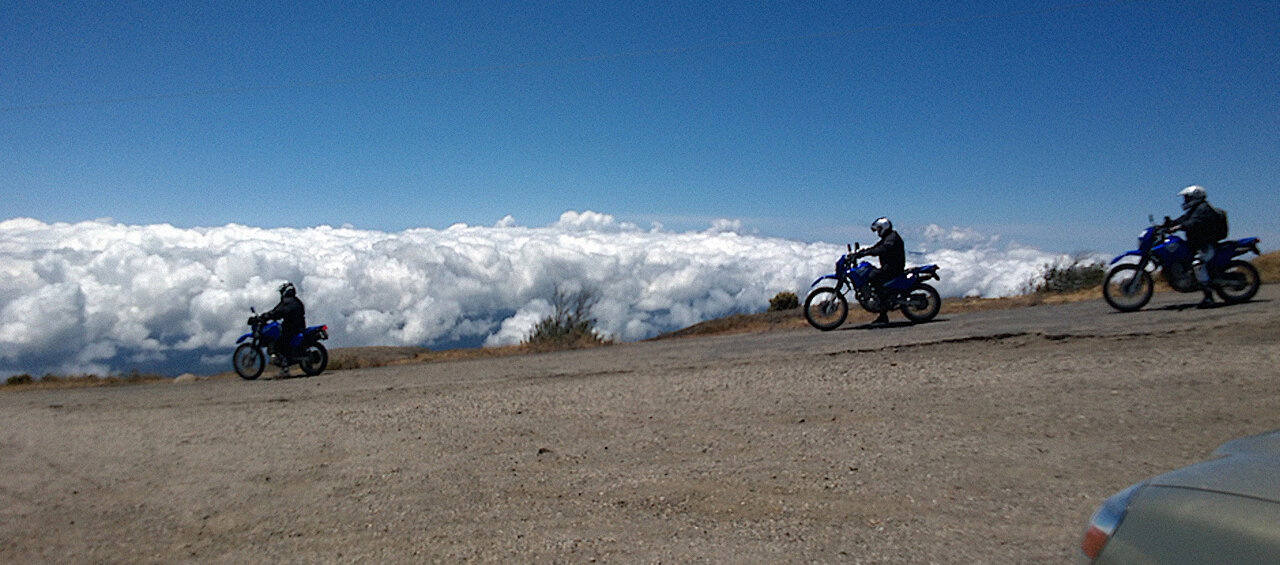 Drei Motorradfahrer fahren auf einer Bergstraße über einem Wolkenmeer