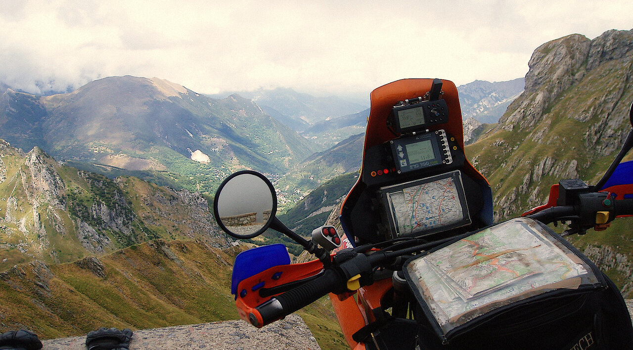 Painel de instrumentos de uma moto com aparelhos de GPS e um mapa de papel, com um vale verde ao fundo