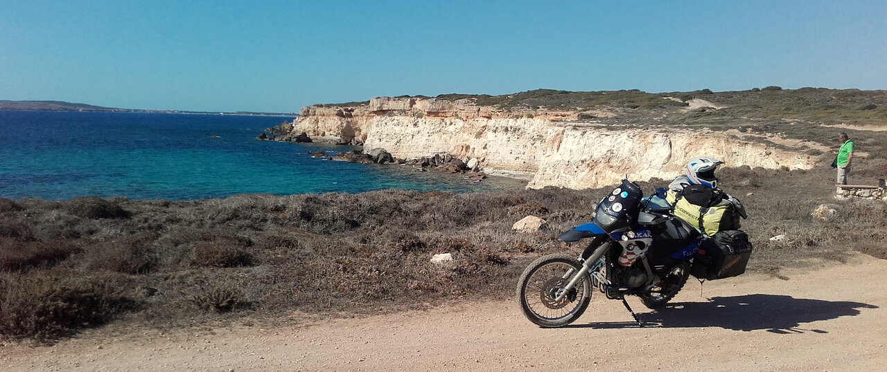 Ein Adventure-Motorrad parkt über türkisfarbenem Wasser und weißen Klippen, mit einem Beobachter in der Nähe der Felsen