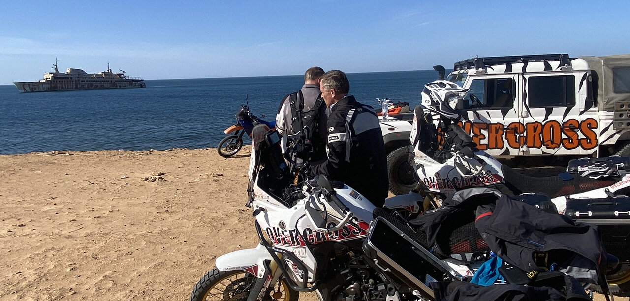 Ein rostendes Schiffswrack vor der Küste nahe einer Klippe, wo Overcross-Motorräder und ein Begleitfahrzeug parken
