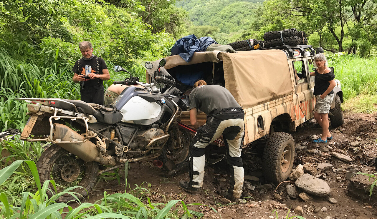 Um camião de expedição 4x4 coberto de lama e uma moto com três viajantes a carregar equipamento na selva densa