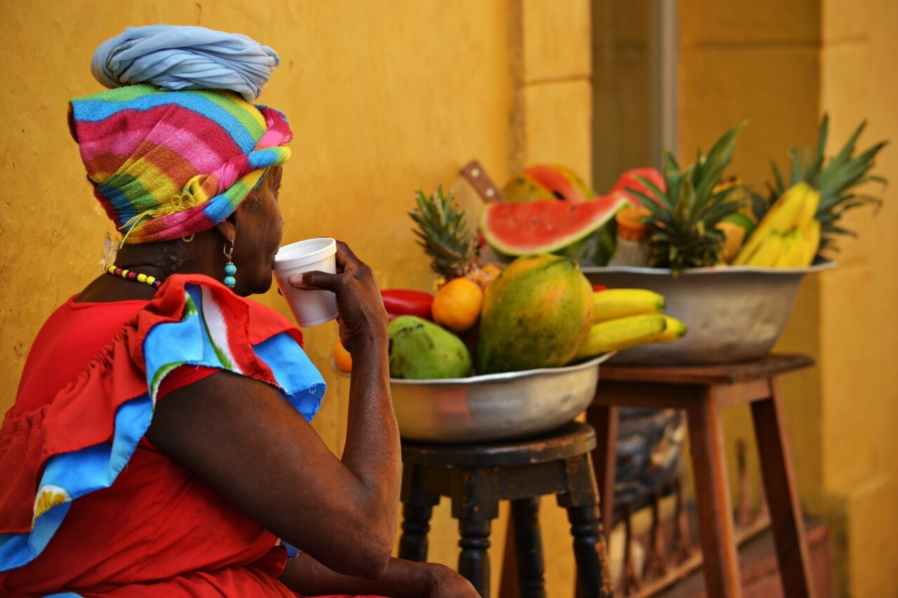 Mulher de turbante colorido e vestido vermelho bebe de uma caneca junto a tigelas de fruta tropical