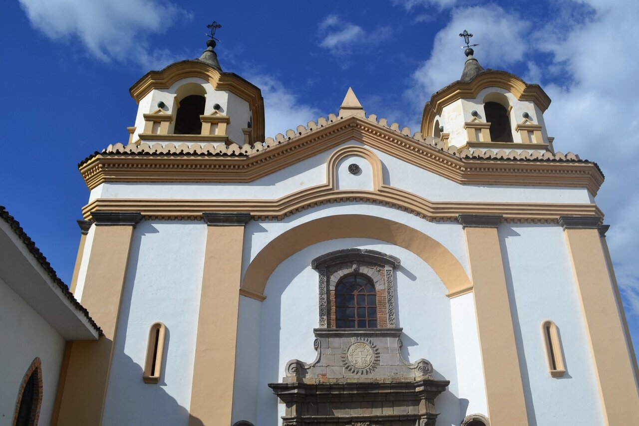 Uma igreja colonial branca com duas torres sineiras e uma entrada em pedra esculpida, sob céu azul