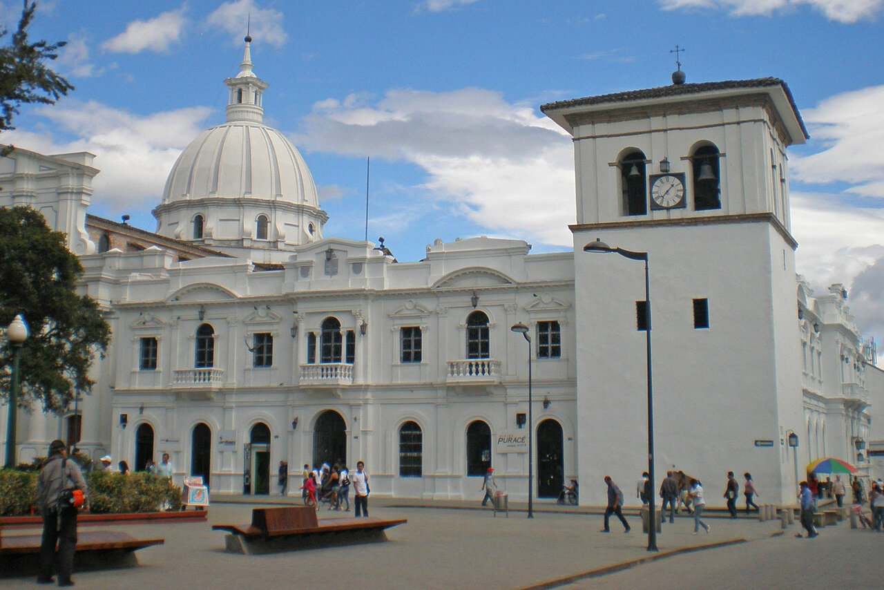 Praça colonial com catedral de cúpula e torre do relógio, pedestres passam por uma placa de loja com o nome Purace