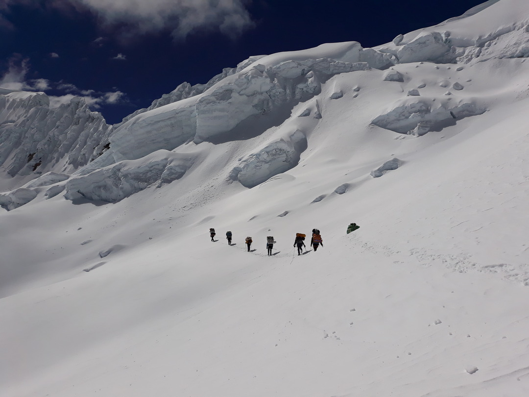 Eine Gruppe von Bergsteigern wandert bei intensivem Sonnenschein und stürmischem Wind zum schneebedeckten Gipfel des Alpamayo in der Cordillera Blanca, Peru