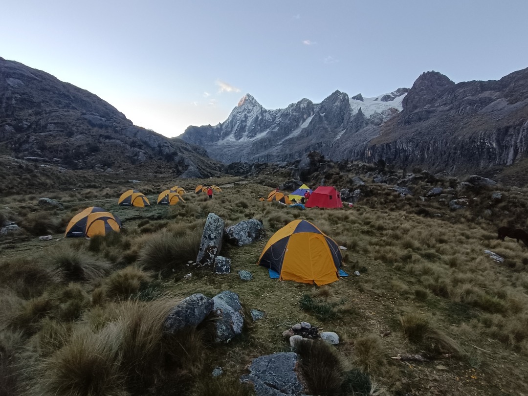 Bergsteiger-Camp mit bunten Zelten in einer Hochgebirgslandschaft der Cordillera Blanca, Peru, mit dem schneebedeckten Alpamayo im Hintergrund bei Sonnenaufgang.