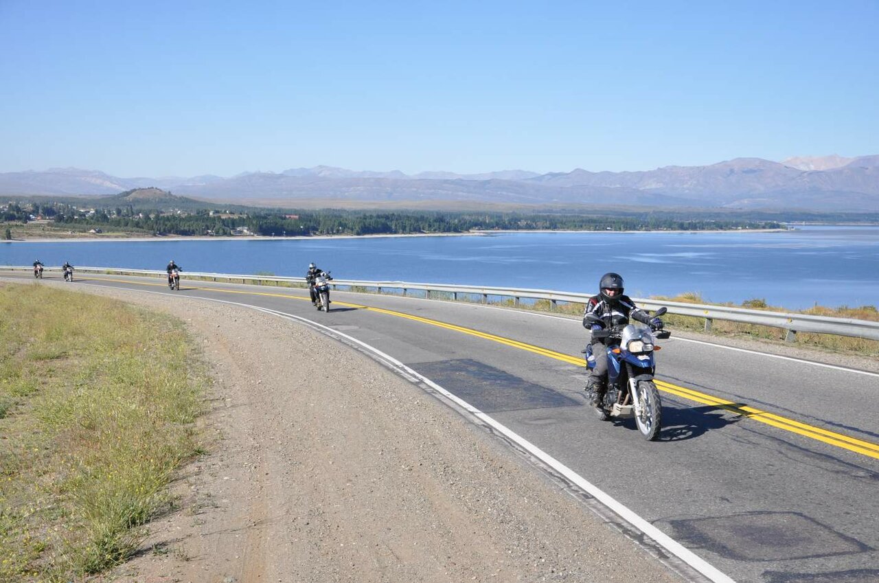Motorradfahrer fahren auf einer asphaltierten Straße neben einem großen blauen See, dahinter Berge