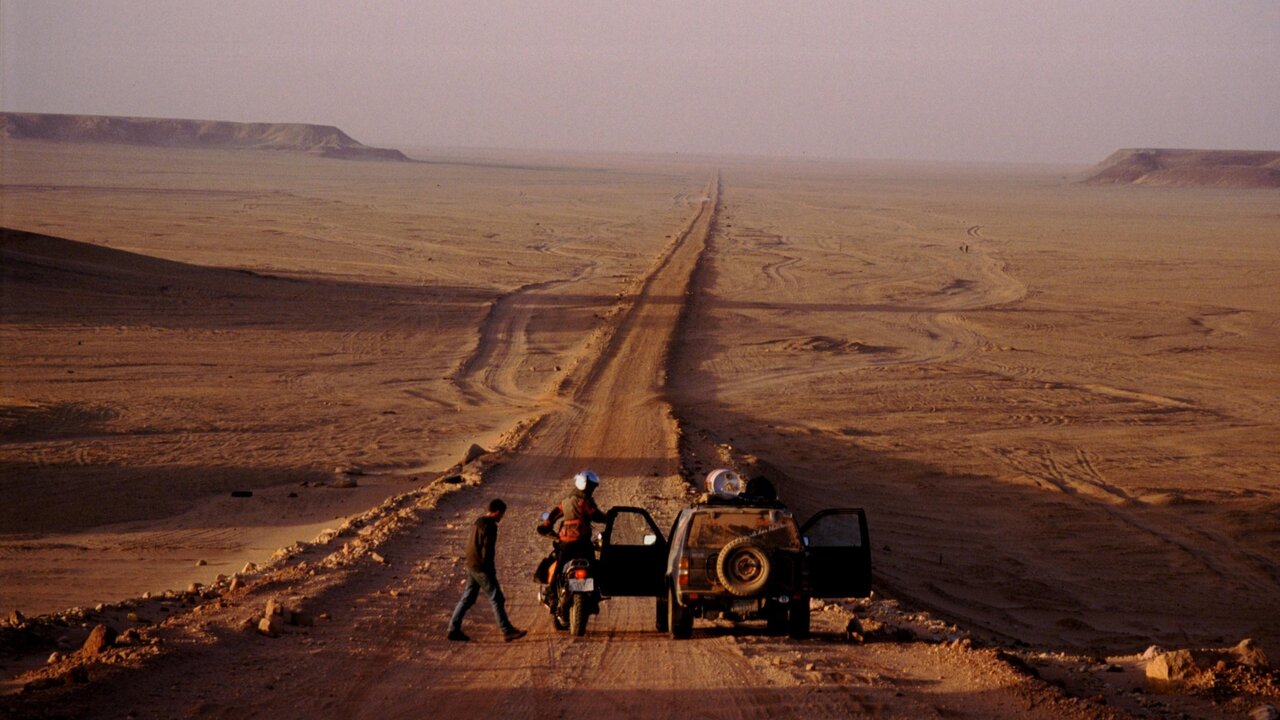 Um motociclista e um veículo de apoio param numa pista no deserto que leva a falésias distantes