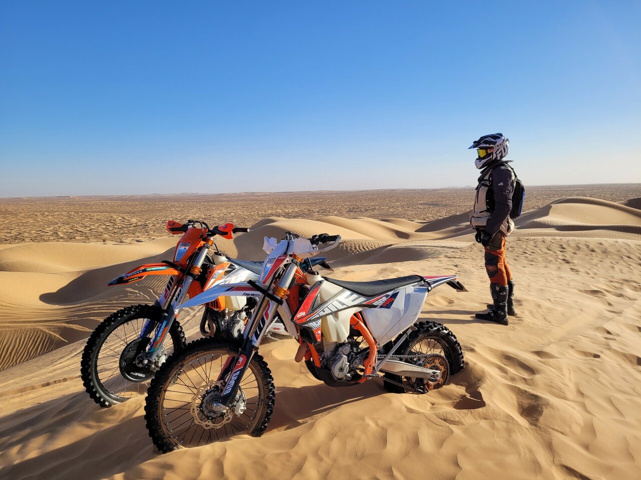 Duas motos de enduro estacionadas numa duna enquanto um piloto observa o deserto