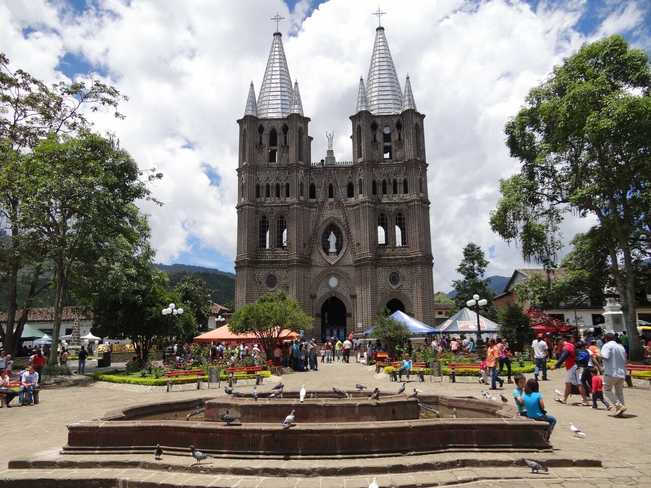 Uma catedral de tijolo com duas torres domina uma praça com pombos e visitantes