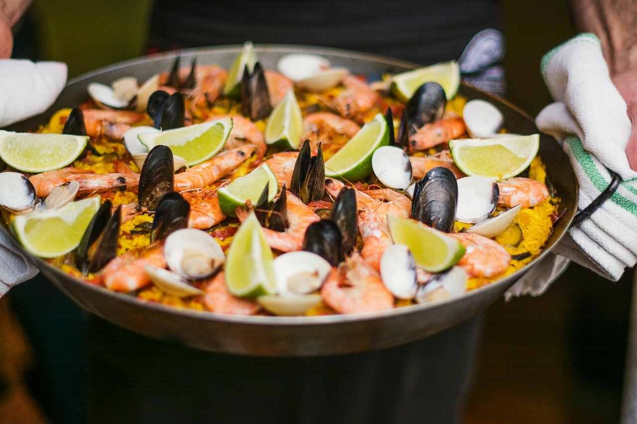 Um grande prato de paella com camarão, mexilhões, amêijoas e gomos de lima, segurado por um empregado