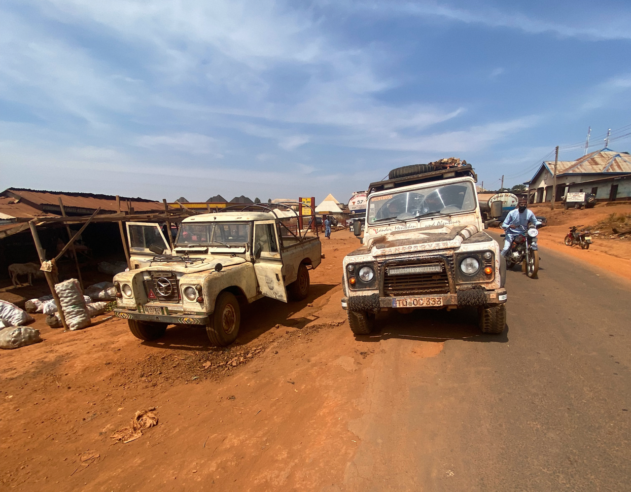 Zwei Land Rover Defender warten in einer staubigen westafrikanischen Marktstraße, während ein Motorradfahrer vorbeifährt