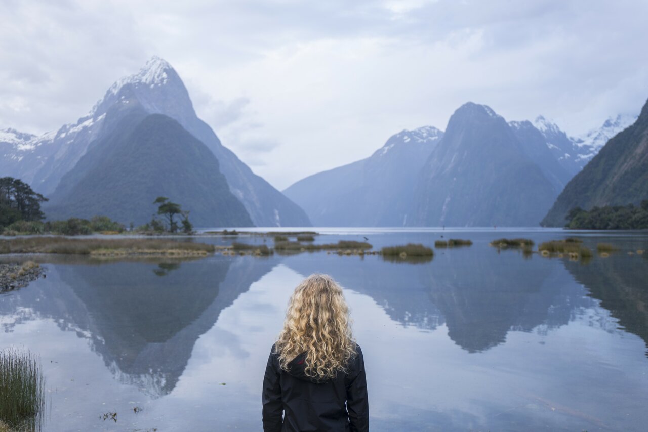 A person stands before a still fjord reflecting steep mountain peaks under a cloudy sky