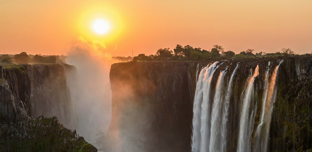 Victoria Falls ao pôr do sol, com luz solar brilhando acima do desfiladeiro enevoado