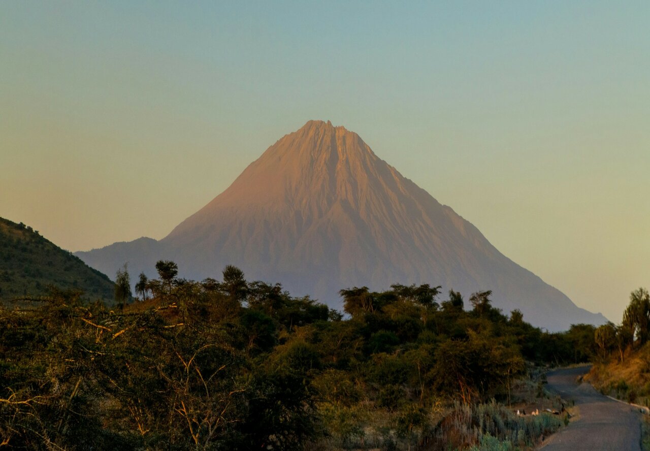 Um pico vulcânico brilha ao pôr do sol sobre vegetação rasteira, com uma estrada de terra em primeiro plano